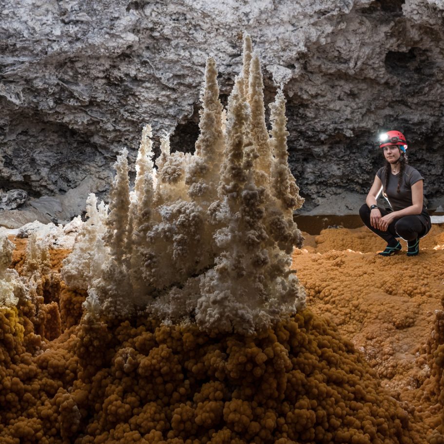 Caver looking at white aragonite "trees" growing on the orange calcite coating of a former cave pool