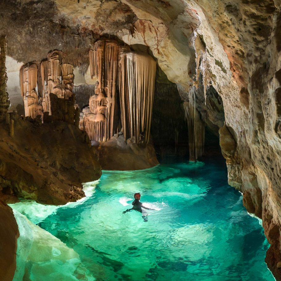 Caver swimming in a large cave pool; flowstone formations in the background