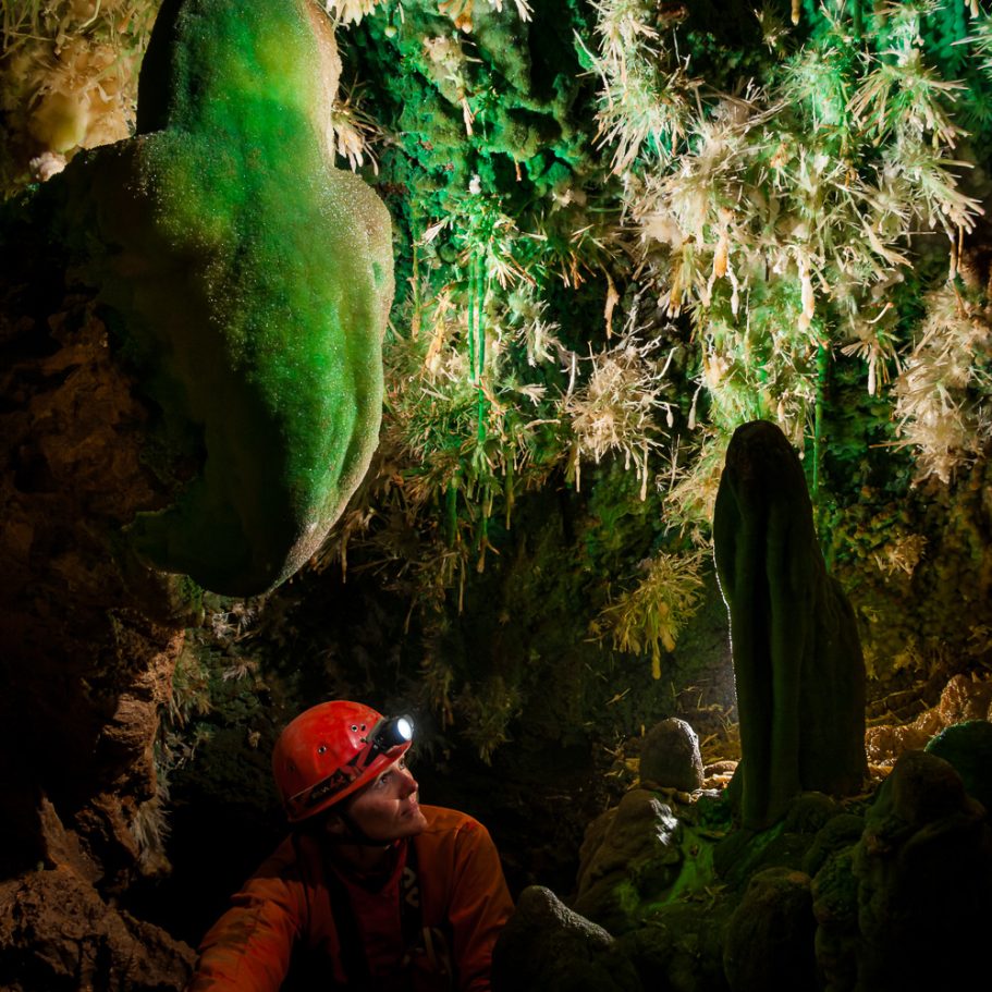 Caver climbing into a littel cave chamber with a profusion of green mineral formations