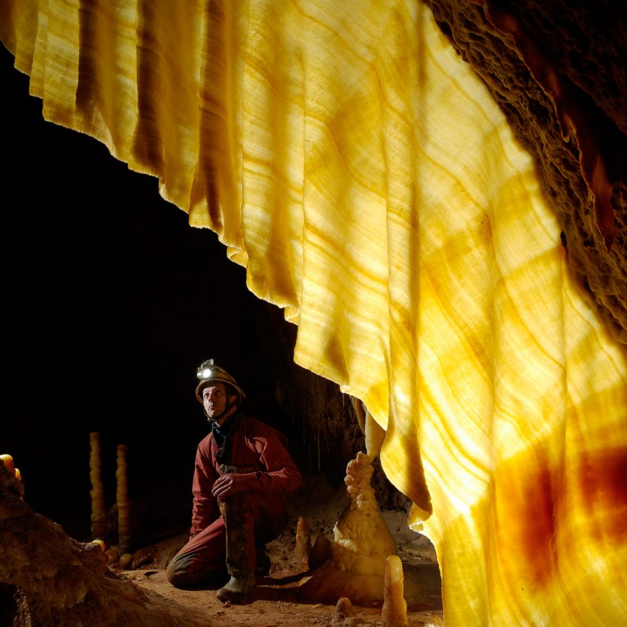 Caver looking at super-delicate and partly translucent calcite drapery "glowing" yellow to orange in backlight