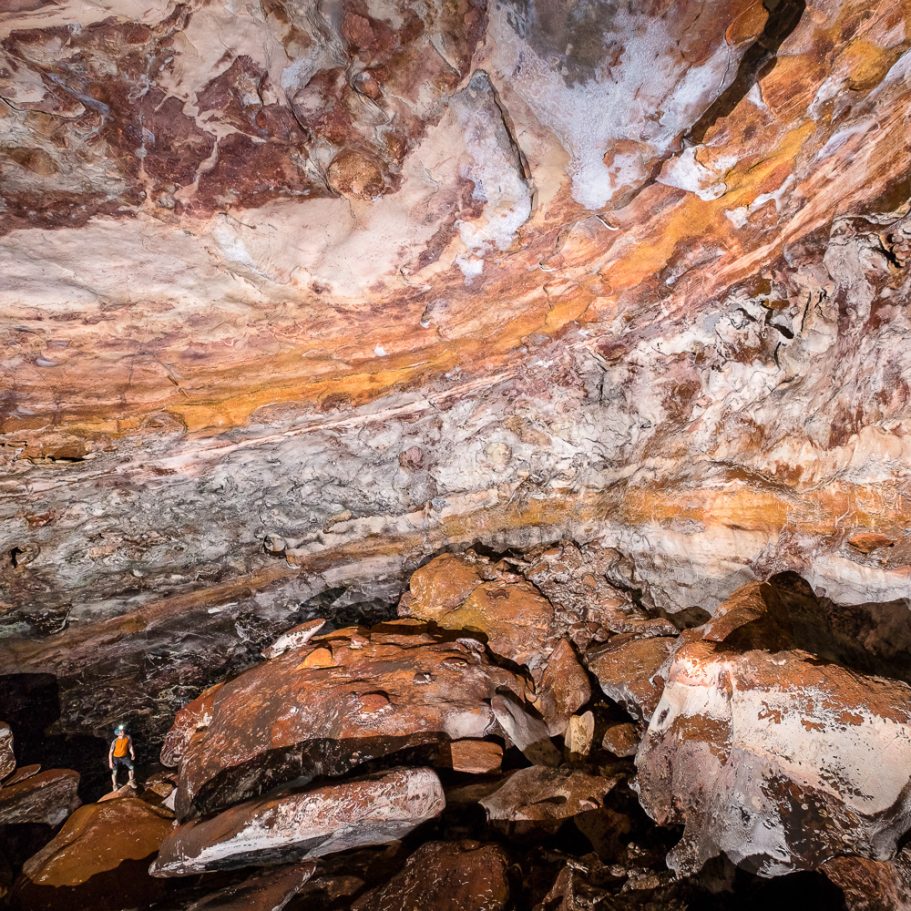 Large cave chamber with walls of orange-brown bedded limestone layers visible in the walls; cave explorer for scale