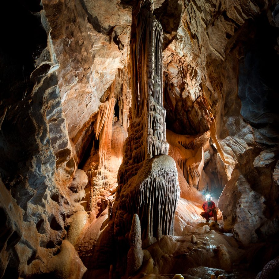 Tall flowstone column in the centre of a cave passage with a caver next to it for scale 