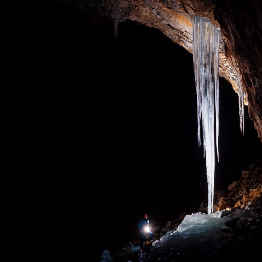 Caver next to a giant ice stalactite in a large cave passage; background pitch black