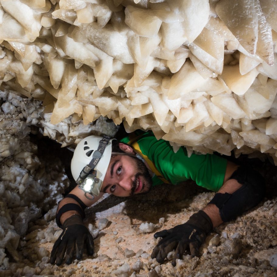 Cave exporer crawling through a low passage with large dogtooth-type calcite spar crystals on the ceiling