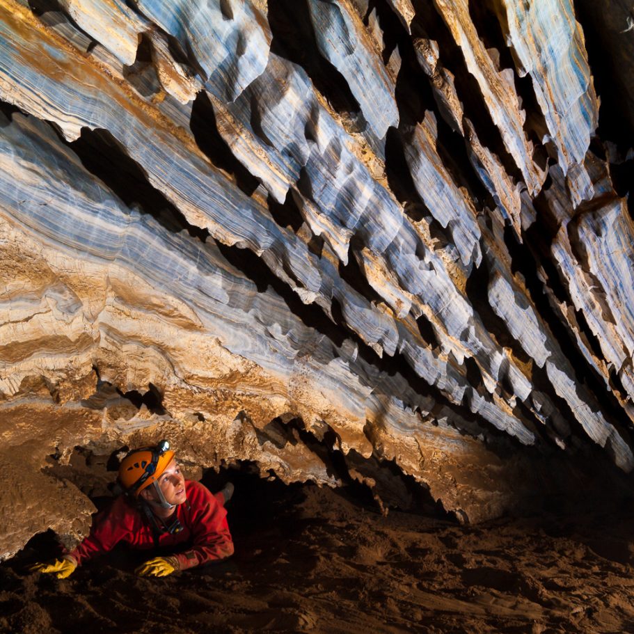 Caver crawling through a low passage with corroded beds of blue marble bedrock